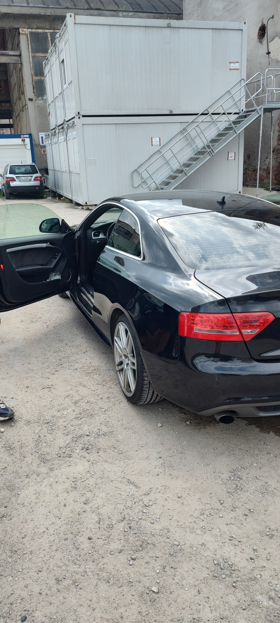 Czarne Audi A5 z otwartymi drzwiami kierowcy, zaparkowane na żwirowym podłożu przed budynkiem kontenerowym z metalowymi schodami.