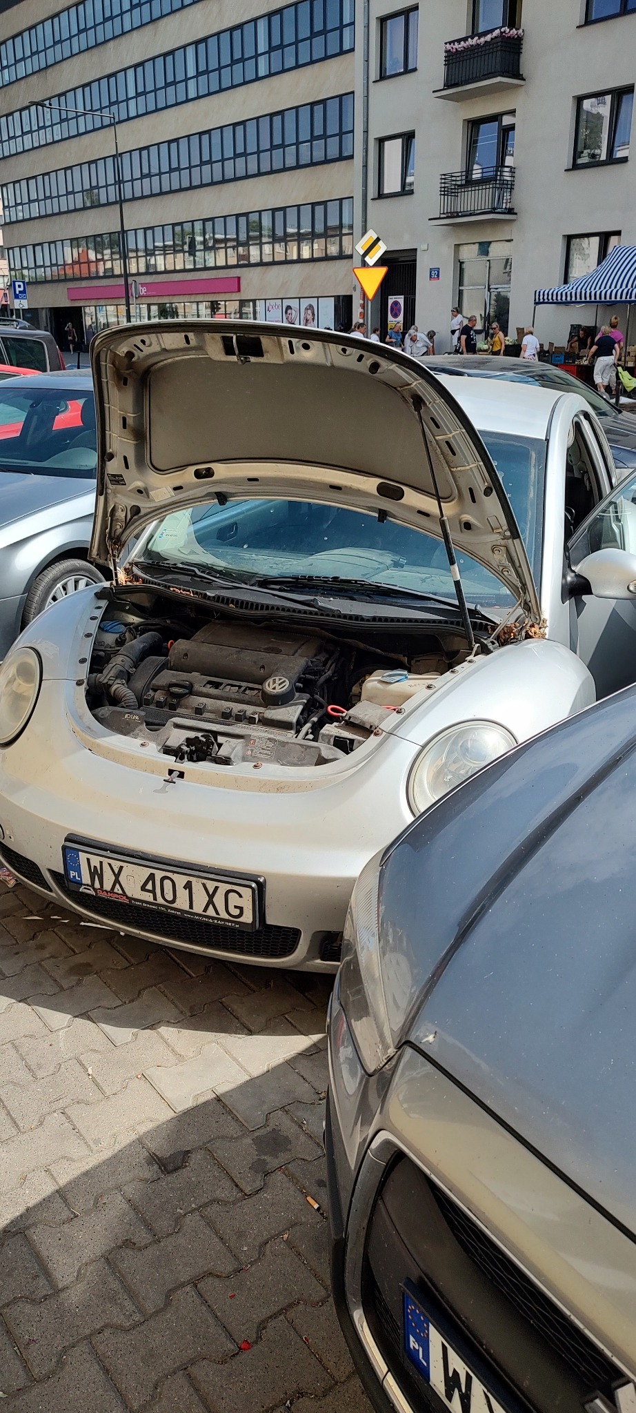 Srebrny Volkswagen Beetle z otwartą maską silnika, zaparkowany na chodniku w miejskiej scenerii, widoczny silnik i tablica rejestracyjna WX 401XG.