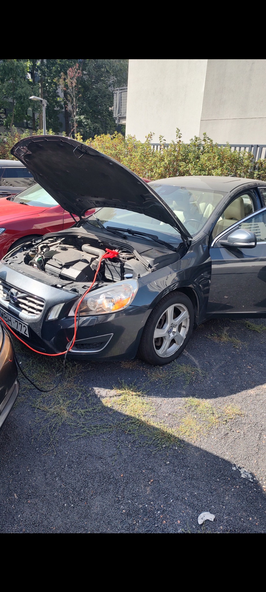 Szary samochód Volvo z otwartą maską, podłączony kablem rozruchowym do akumulatora innego pojazdu, stojący na żwirowym parkingu w słoneczny dzień.