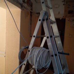 Marcin Włodarczyk Diagnosis - Aluminiowa drabina rozstawiona w remontowanym pomieszczeniu z odsłoniętą cegłą, na której zawieszony jest zwój kabli elektrycznych w białej izolacji.