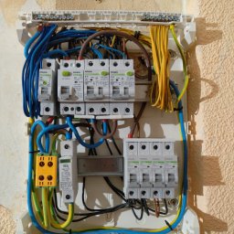 Marcin Włodarczyk Diagnosis - Otwarta skrzynka bezpiecznikowa z widocznymi bezpiecznikami, przewodami w kolorach niebieskim, żółtym, zielonym i brązowym, zamontowana na nierównej, pomalowanej ścianie.