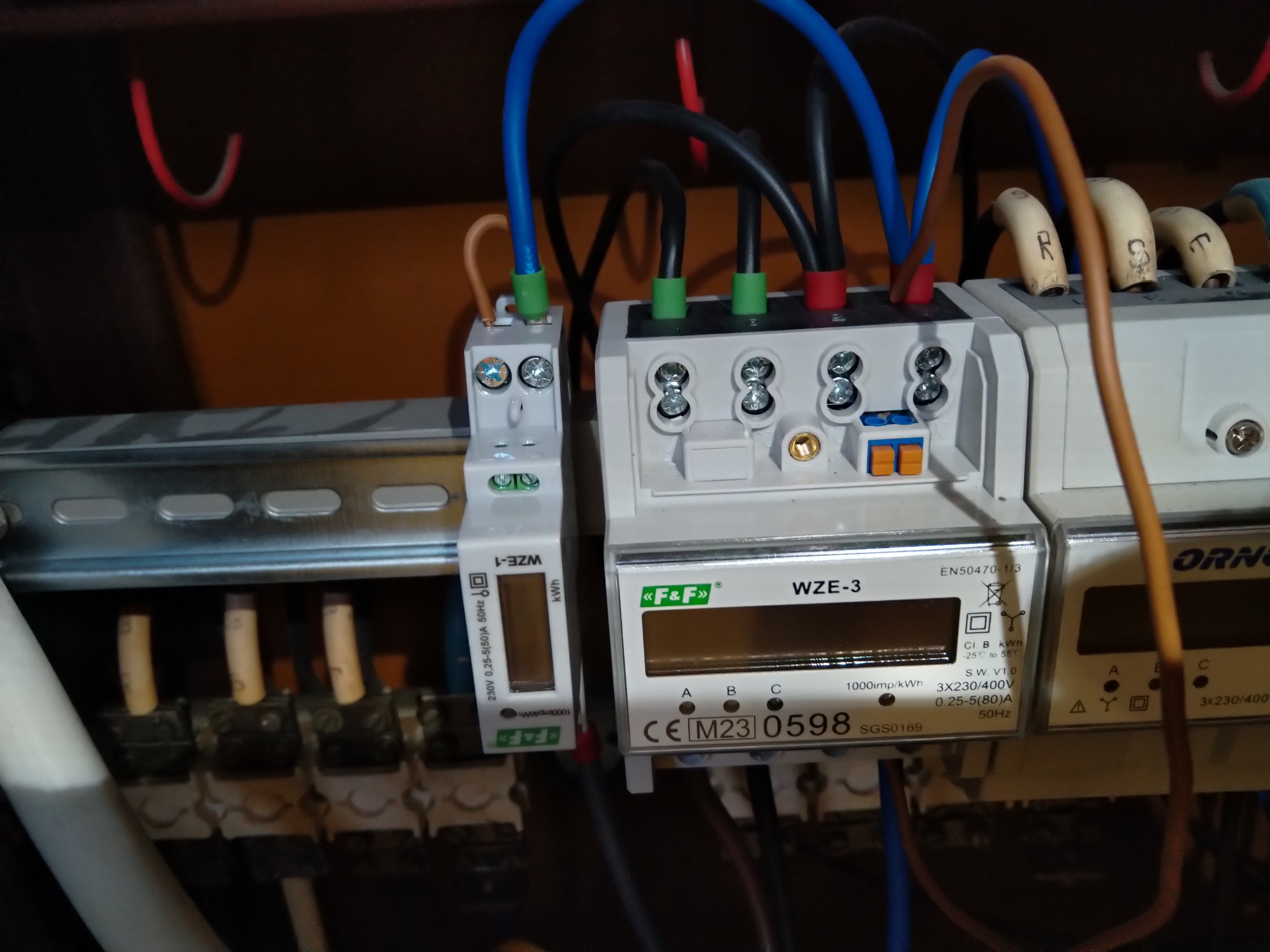 Szczegółowe ujęcie zamontowanych liczników energii elektrycznej WZE-1 i WZE-3 firmy F&F z podłączonymi przewodami w kolorach niebieskim, czarnym, czerwonym i brązowym, zamocowanych na szynie DIN...