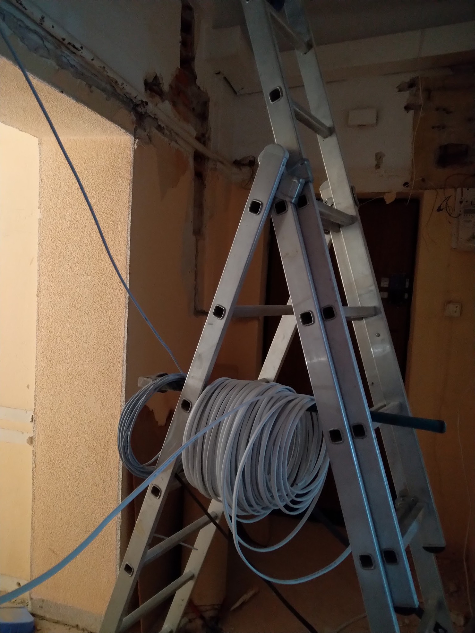 Aluminiowa drabina rozstawiona w remontowanym pomieszczeniu z odsłoniętą cegłą, na której zawieszony jest zwój kabli elektrycznych w białej izolacji.