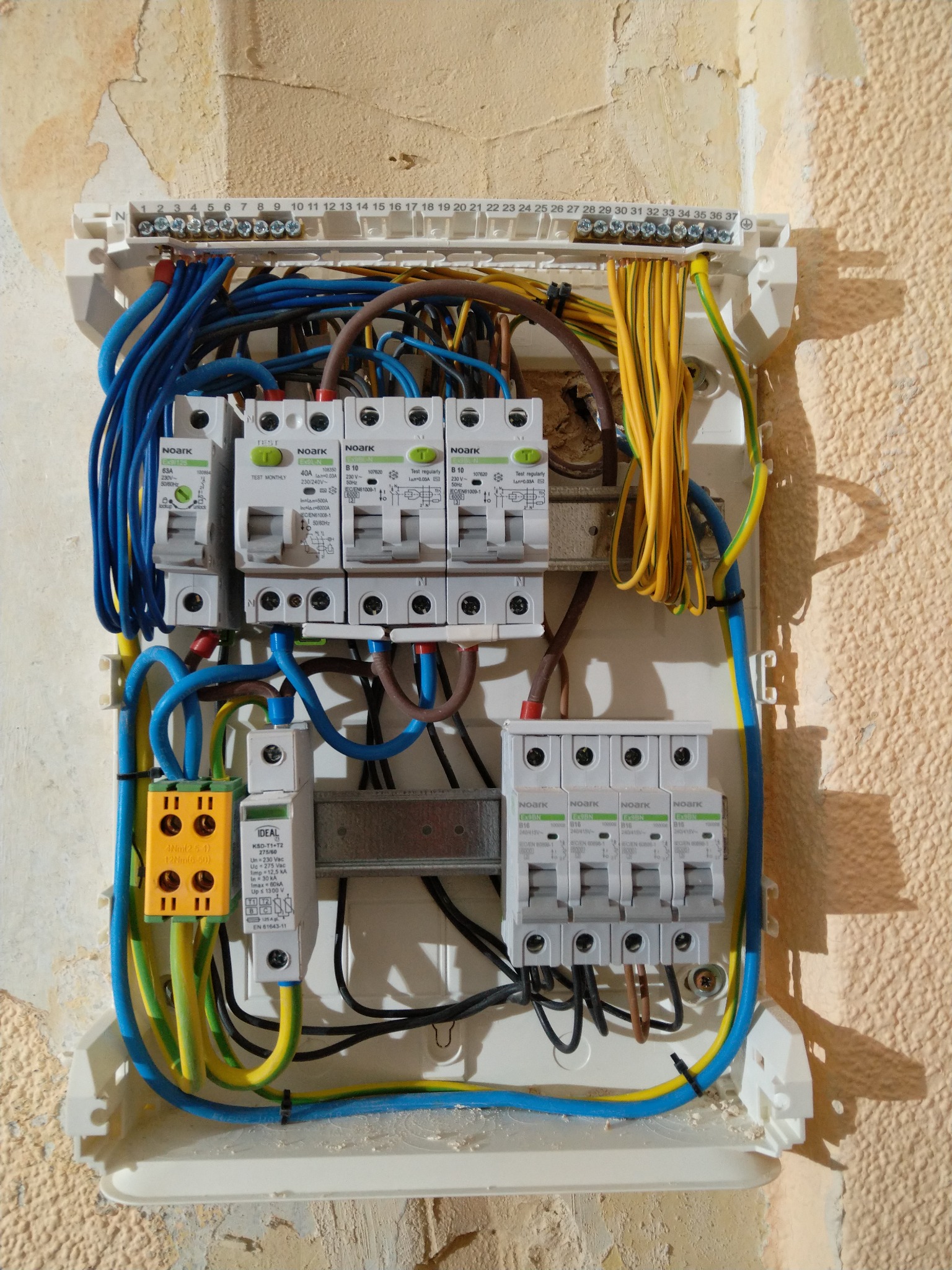 Otwarta skrzynka bezpiecznikowa z widocznymi bezpiecznikami, przewodami w kolorach niebieskim, żółtym, zielonym i brązowym, zamontowana na nierównej, pomalowanej ścianie.