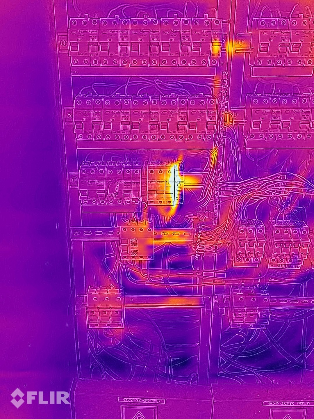Termowizyjny obraz rozdzielnicy elektrycznej, ukazujący punktowe przegrzanie jednego z wyłączników, z widocznymi przewodami i elementami zabezpieczeń.
