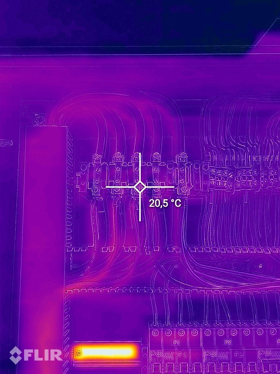 Termowizja rozdzielnicy elektrycznej, widoczne kable i bezpieczniki, wskazanie temperatury 20,5 stopni Celsjusza, obraz w odcieniach fioletu i żółci.