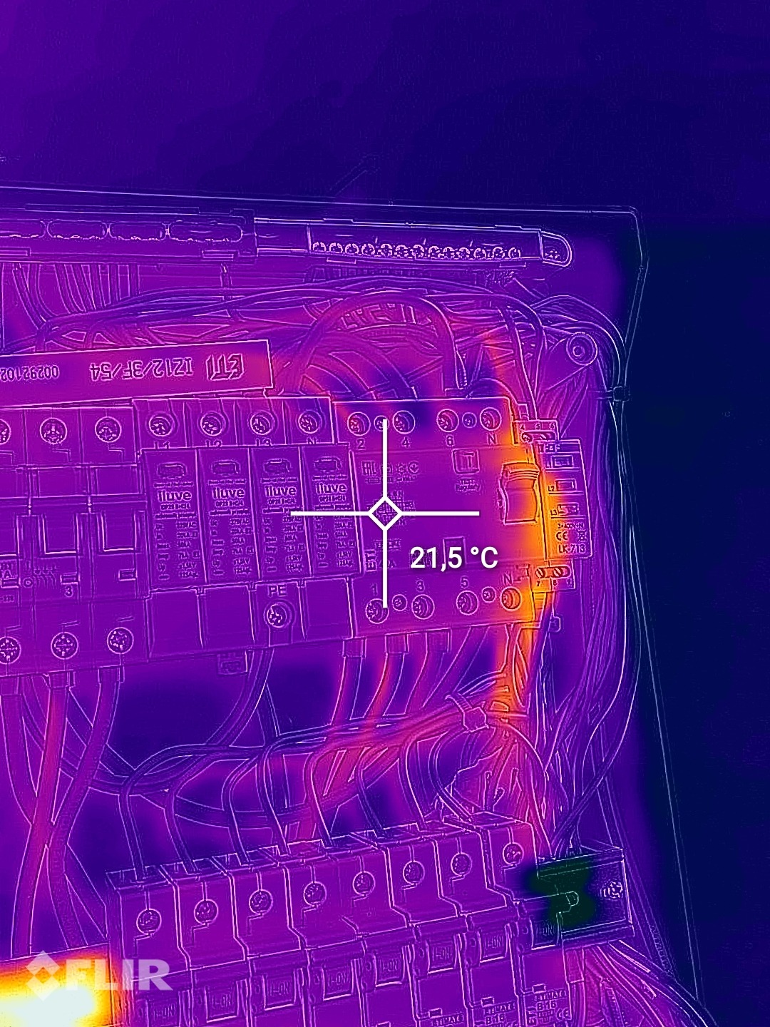Termowizyjne zdjęcie skrzynki elektrycznej z widocznymi przewodami i bezpiecznikami, wskazujące temperaturę 21,5 stopni Celsjusza w jednym z punktów.