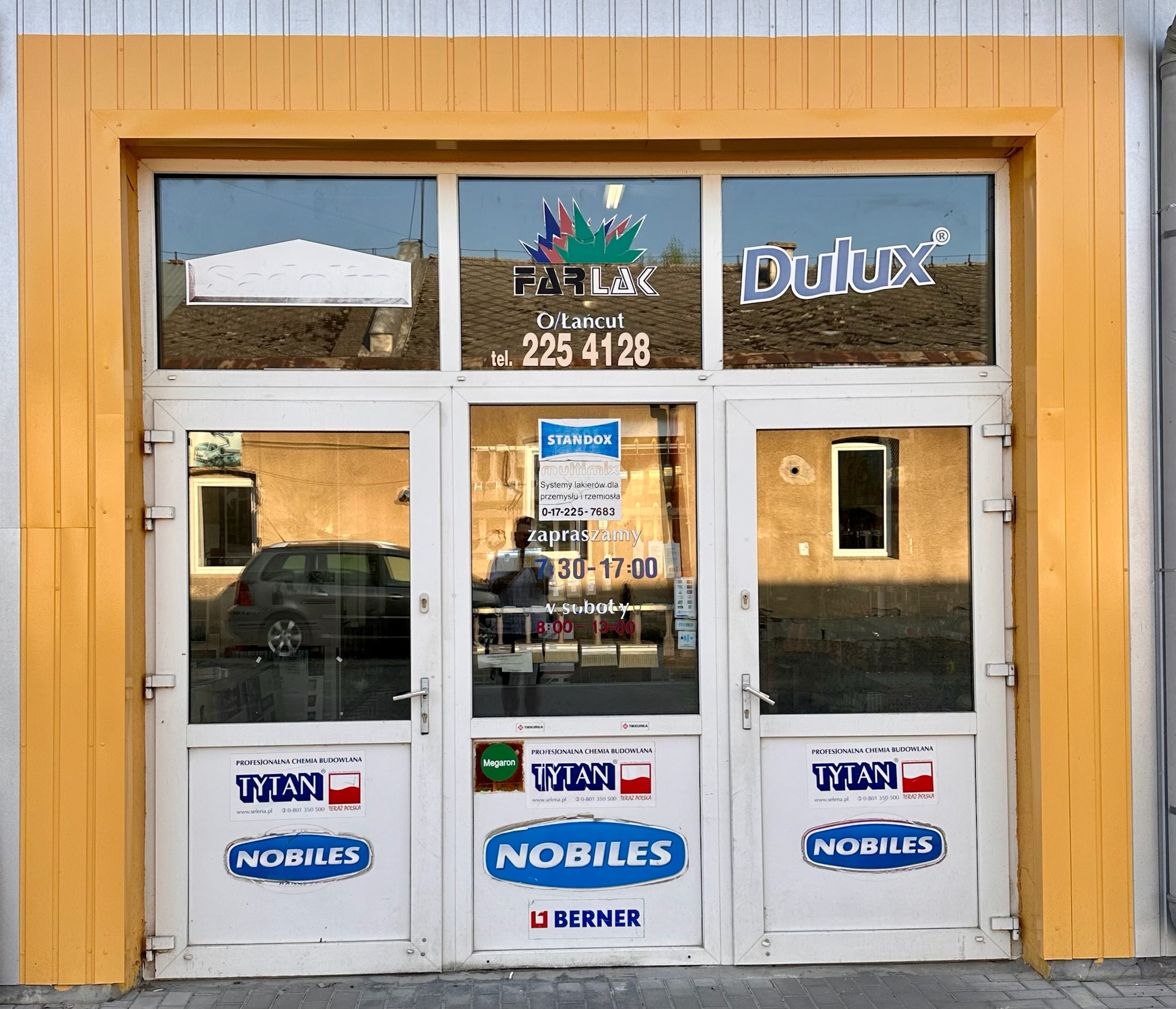 Fasada sklepu z farbami, na szybach naklejone reklamy marek Farlak, Dulux, Standox, na drzwiach naklejki Tytan i Nobiles, żółte obramowanie.