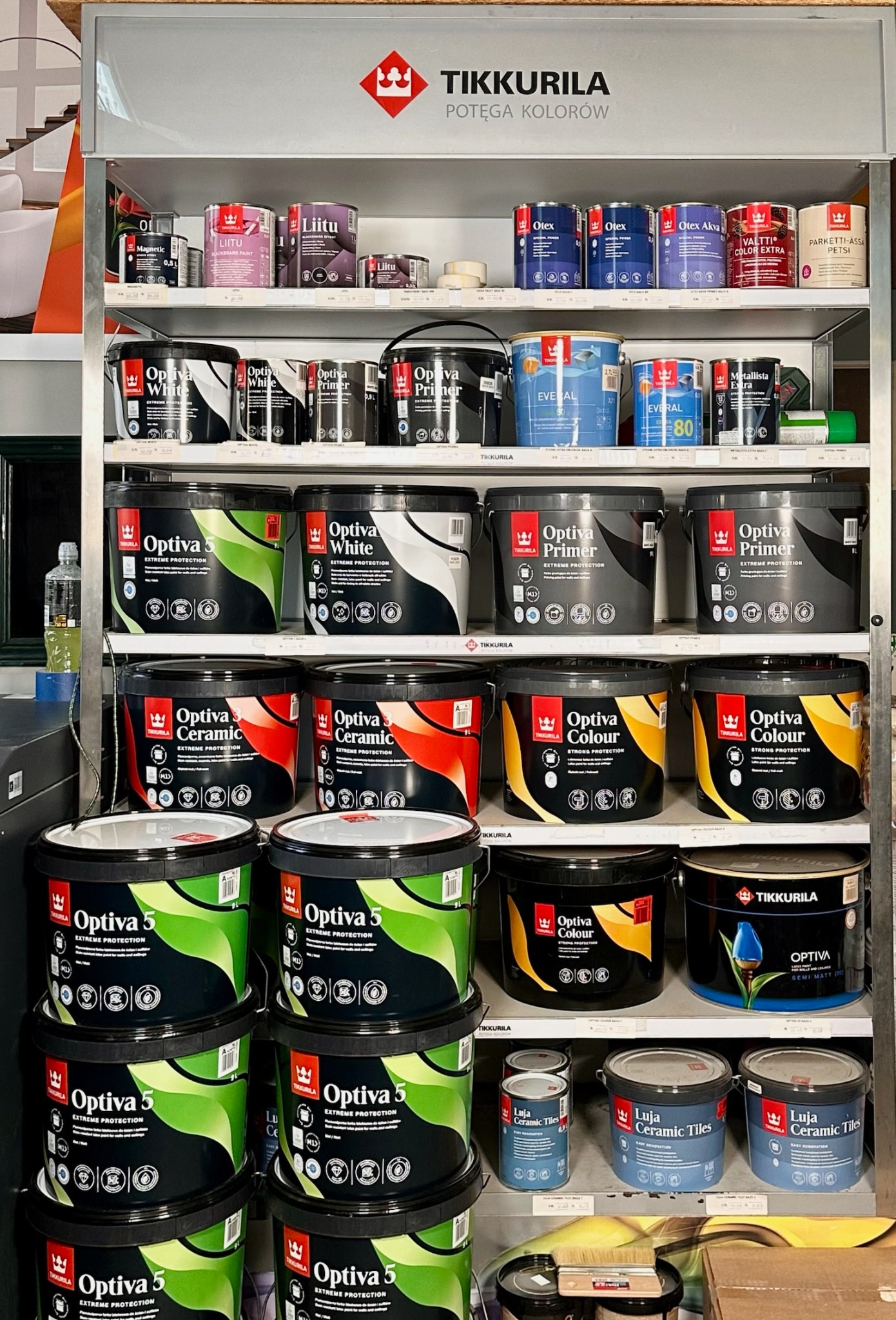 Półki w sklepie z farbami Tikkurila, prezentujące różne kolory i rodzaje farb, w tym Optiva 5, Optiva White, Optiva Primer, Optiva Ceramic, Optiva Colour oraz Luja Ceramic Tiles.