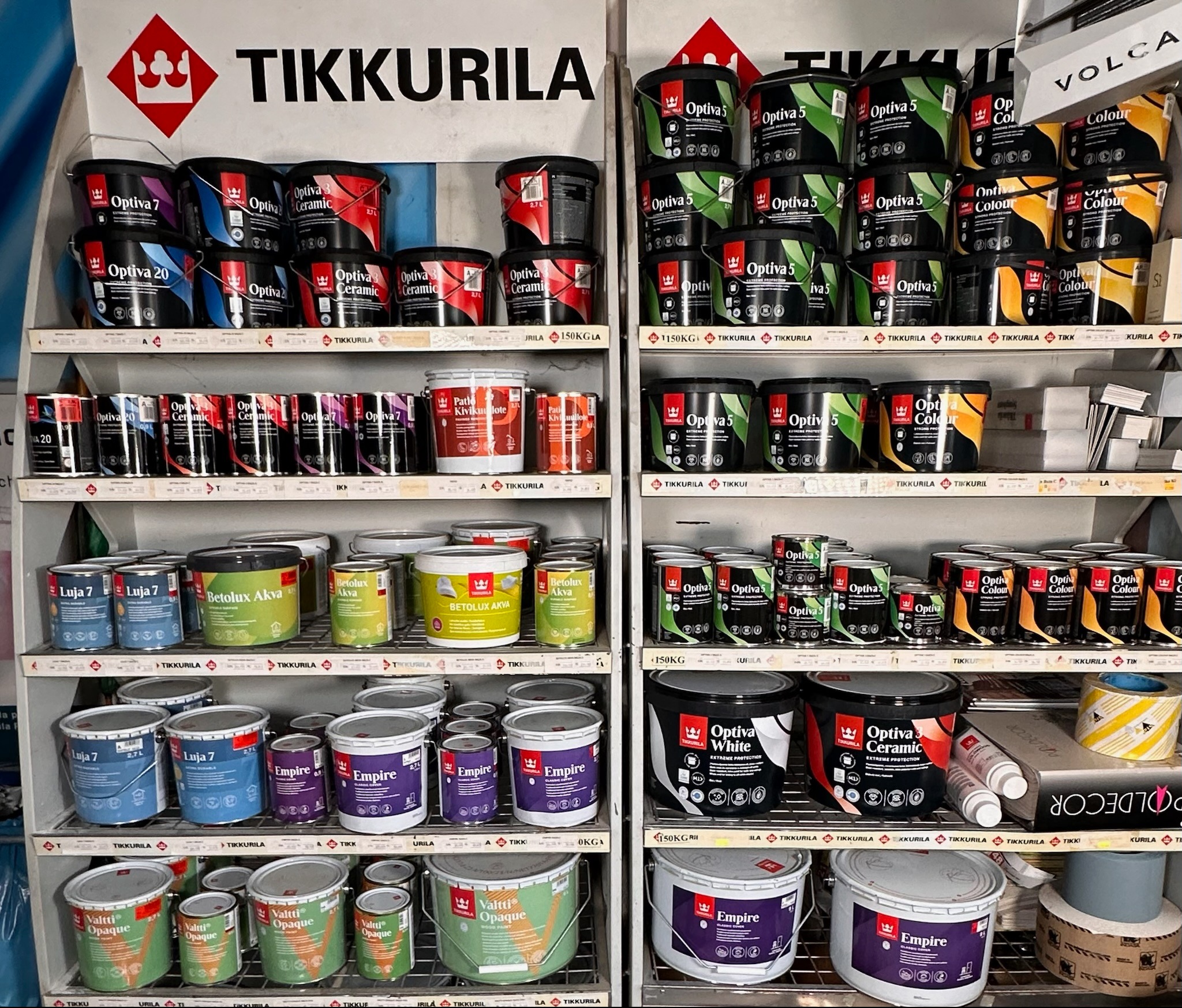 Półki w sklepie z farbami Tikkurila, wypełnione różnymi rodzajami farb w puszkach, w tym Optiva, Luja, Betolux Akva, Valtti Opaque i Empire, ułożone kolorystycznie.