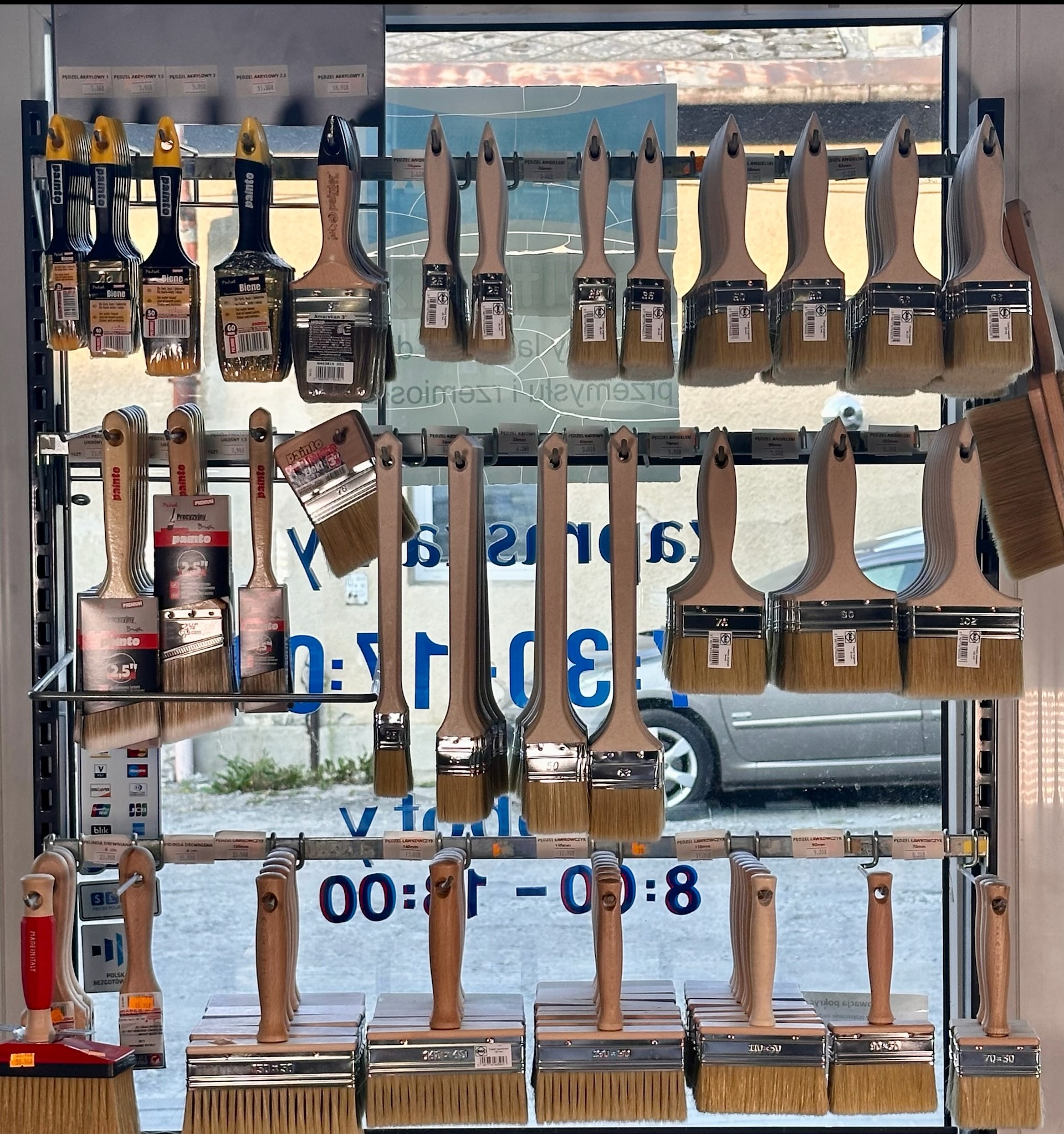 Ekspozycja różnych pędzli malarskich na metalowym regale w sklepie, widoczne pędzle akrylowe, angielskie, ławkowce i inne, niektóre z etykietami cenowymi, na tle okna z częściowo widocznym napisem...