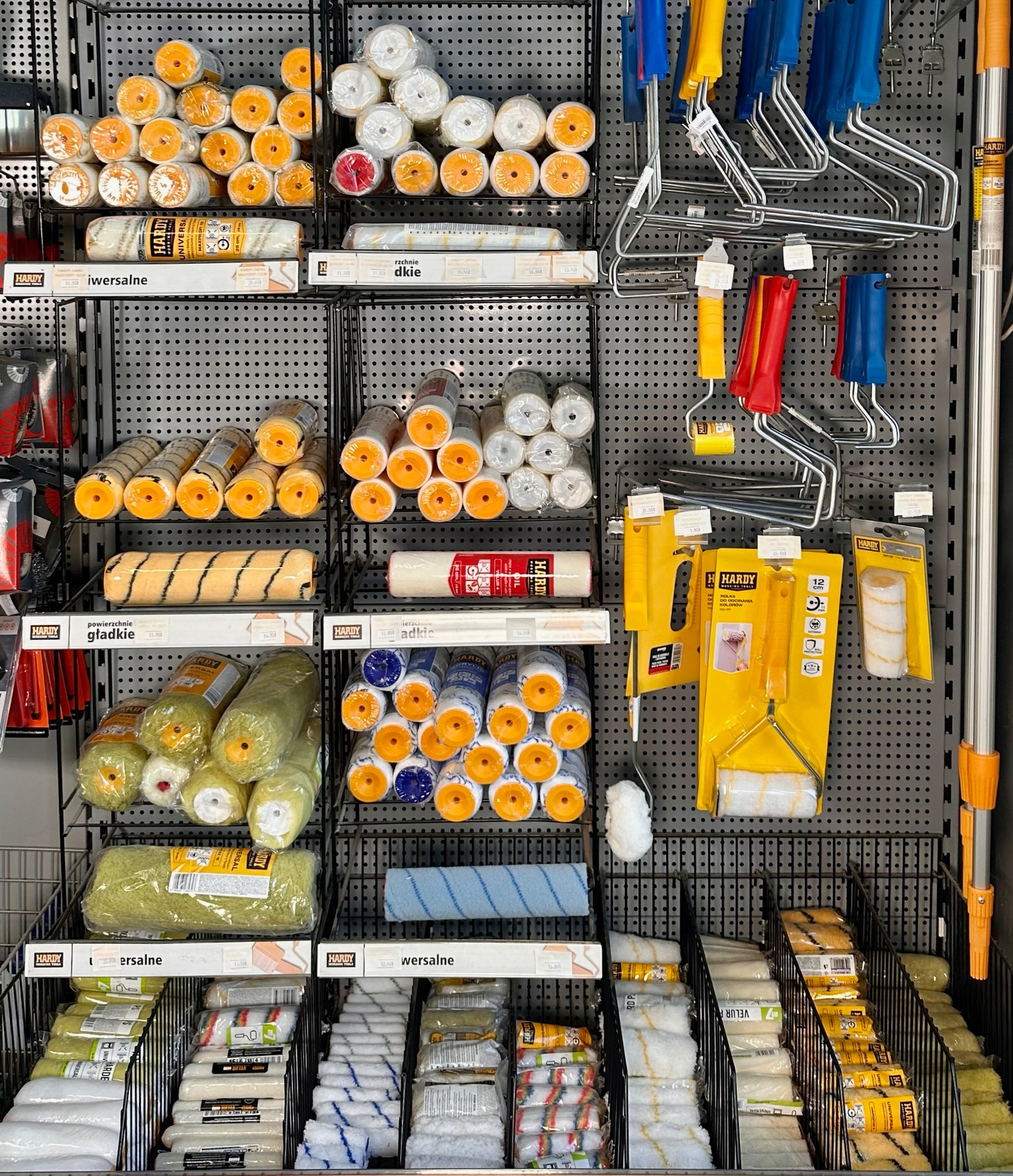 Ekspozycja w sklepie budowlanym: Różne rodzaje wałków malarskich Hardy, uchwyty do wałków i teleskopowe przedłużki na perforowanej ścianie.