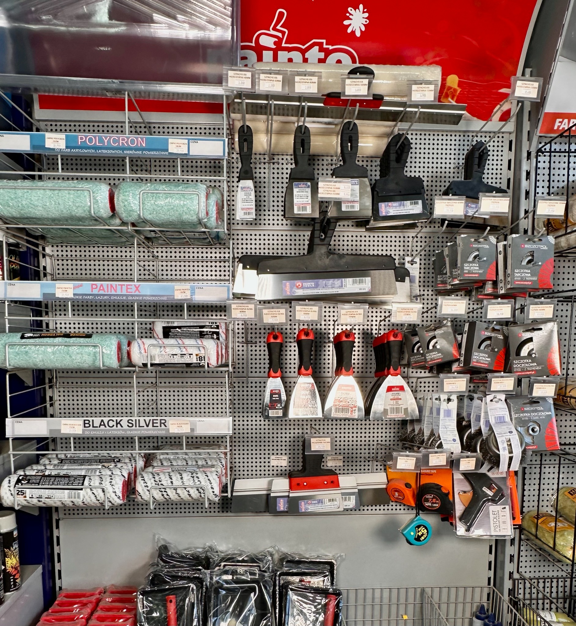 Ekspozycja w sklepie budowlanym: wałki malarskie różnych typów (Polycron, Paintex, Black Silver) obok szpachelek z czerwonymi uchwytami, szczotek drucianych i narzędzi pomiarowych (miarki zwijane).