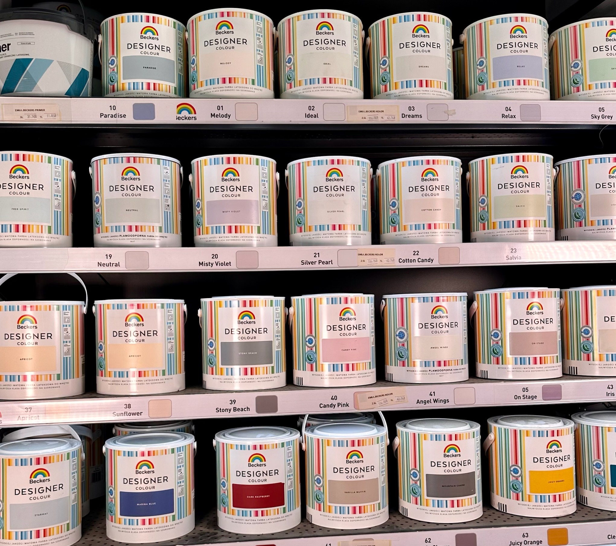 Różnokolorowe puszki farb Beckers Designer Colour na półce sklepowej, z widocznymi nazwami odcieni jak Paradise, Melody, Ideal, Dreams, Sky Grey, Neutral, Misty Violet, Cotton Candy, Apricot...