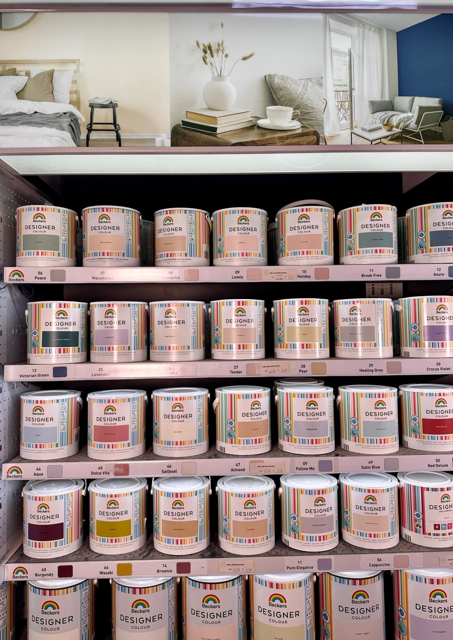 Półka w sklepie z farbami Beckers Designer Colour, prezentująca szeroką gamę odcieni w puszkach, z aranżacjami wnętrz w tle, ilustrującymi potencjalne efekty kolorystyczne.