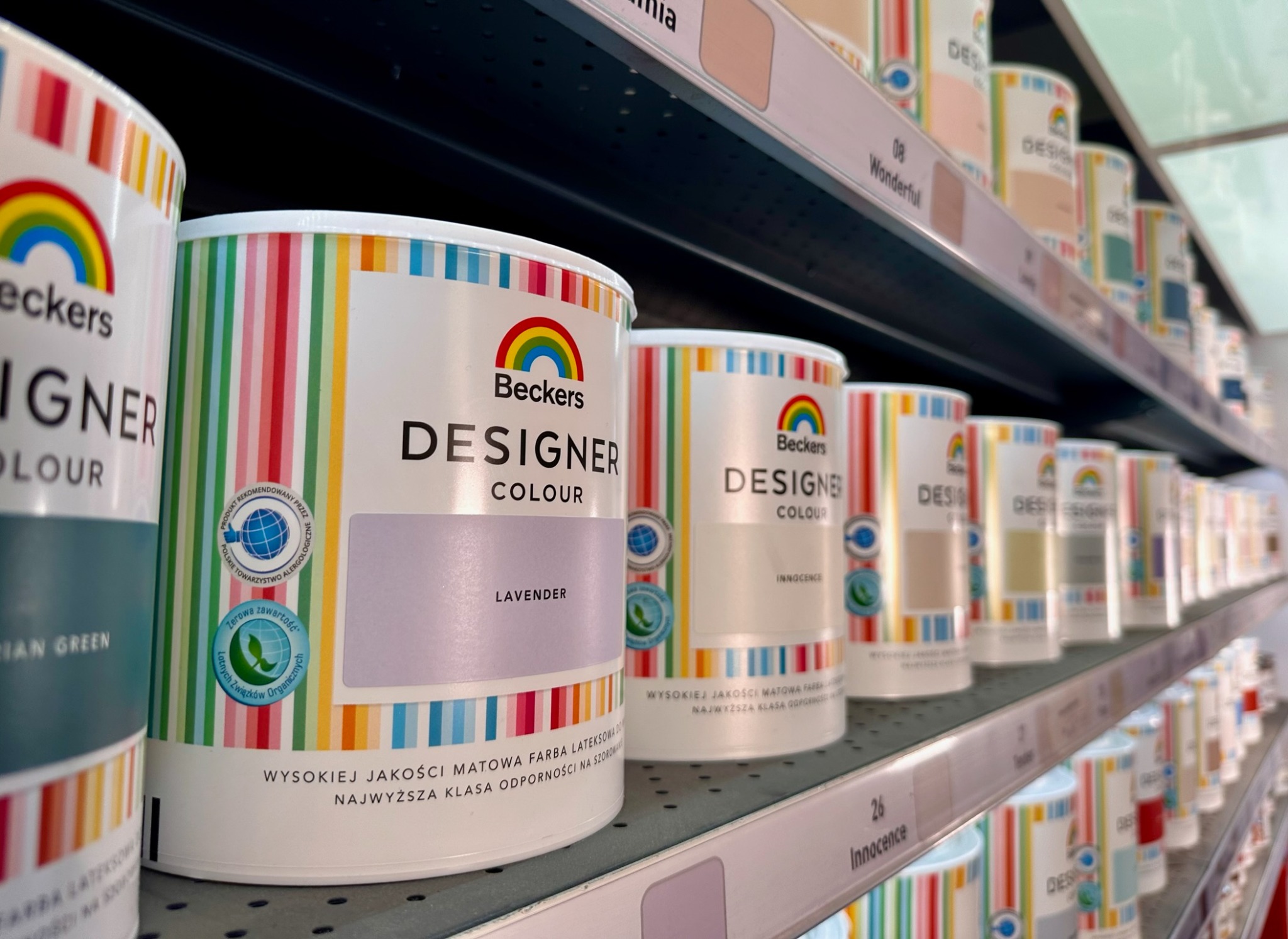Półka sklepowa z puszkami farb Beckers Designer Colour w odcieniach Lavender, Innocence i innych, z etykietami informującymi o wysokiej jakości i rekomendacji Polskiego Towarzystwa Alergologicznego.