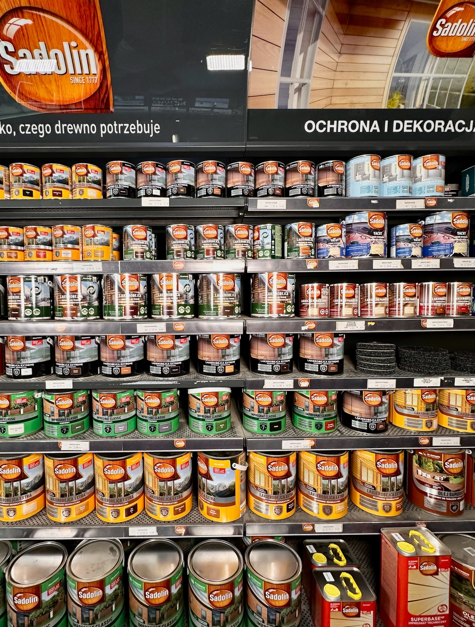 Półki sklepowe wypełnione puszkami z lakierobejcami i impregnatami do drewna marki Sadolin, różne kolory i rodzaje, widoczny napis 'Ochrona i dekoracja'.