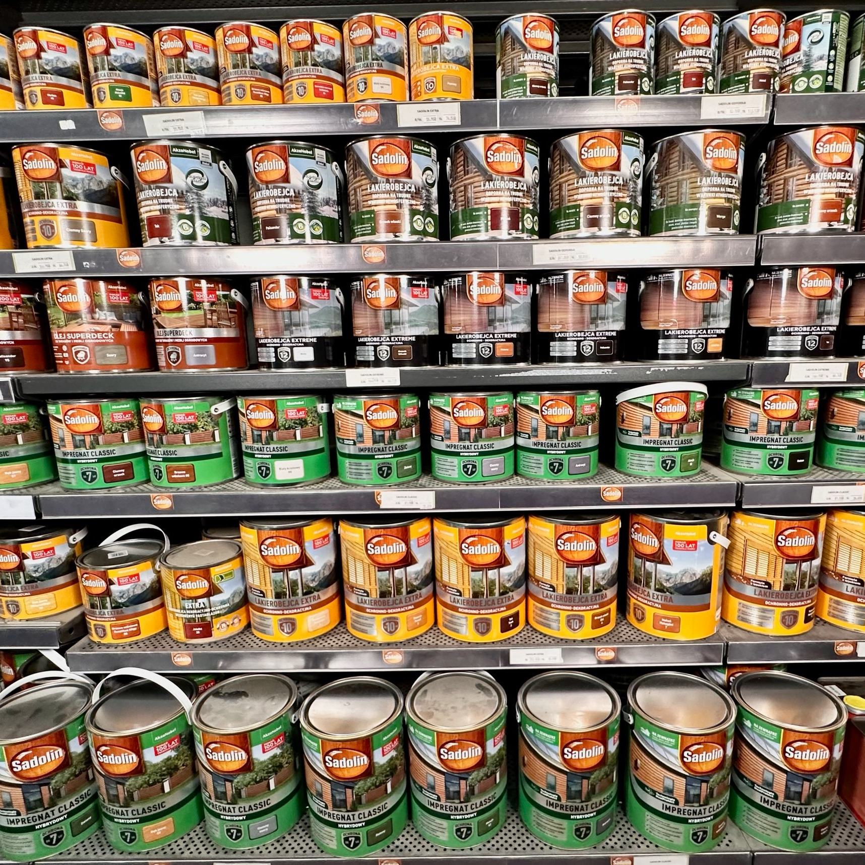 Półki w sklepie z farbami i lakierami do drewna marki Sadolin, różne rodzaje i kolory impregnatów i lakierobejc, widok z bliska.