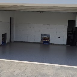 Elpox - Wykończony garaż z szarą posadzką z delikatnym wzorem, białe płytki na ścianach, widoczne instalacje i osprzęt elektryczny, jasne wnętrze.