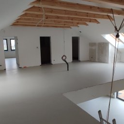 Elpox - Jasnoszara, gładka podłoga w przestronnym, surowym wnętrzu poddasza z drewnianymi belkami stropowymi i pionowymi słupami, widoczne okno dachowe i otwory drzwiowe.