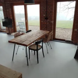 Jasnoszara podłoga w przestronnym wnętrzu z ceglanymi ścianami, drewnianym stołem i widokiem na ogród przez duże okna.