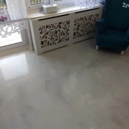 Jasnoszara, błyszcząca posadzka w pokoju z turkusowym fotelem i białym grzejnikiem z dekoracyjnym ażurem.