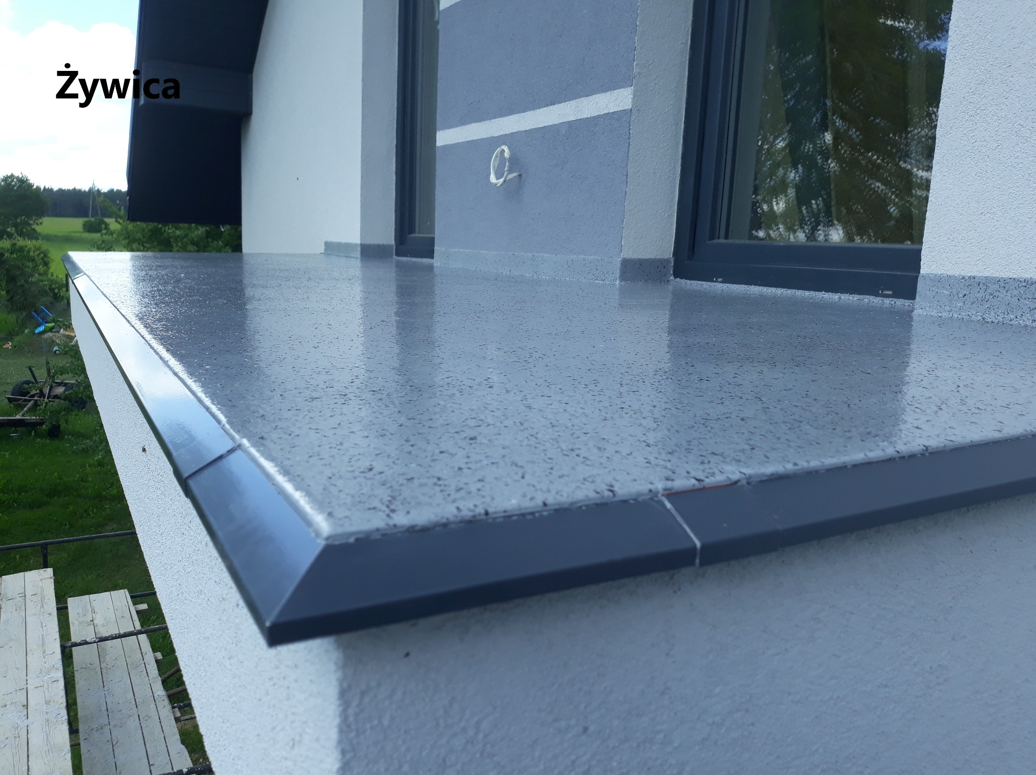 Balkon z szarą, błyszczącą powierzchnią z drobnymi ciemnymi plamkami, zakończony ciemną listwą, widok z dołu na fragment elewacji i okno.