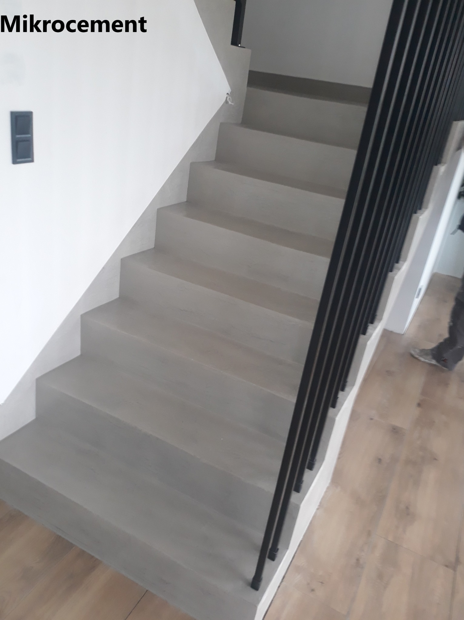 Schody wewnętrzne pokryte szarą, gładką powłoką z czarną metalową balustradą, widoczne z dołu, z fragmentem drewnianej podłogi w tle.