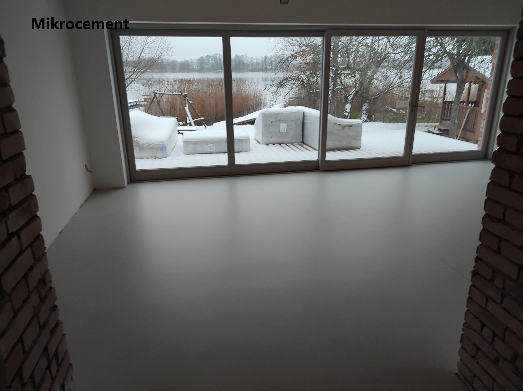 Jasnoszara, gładka podłoga w nowym pomieszczeniu z dużym oknem tarasowym wychodzącym na zasypany śniegiem ogród z jeziorem w tle.