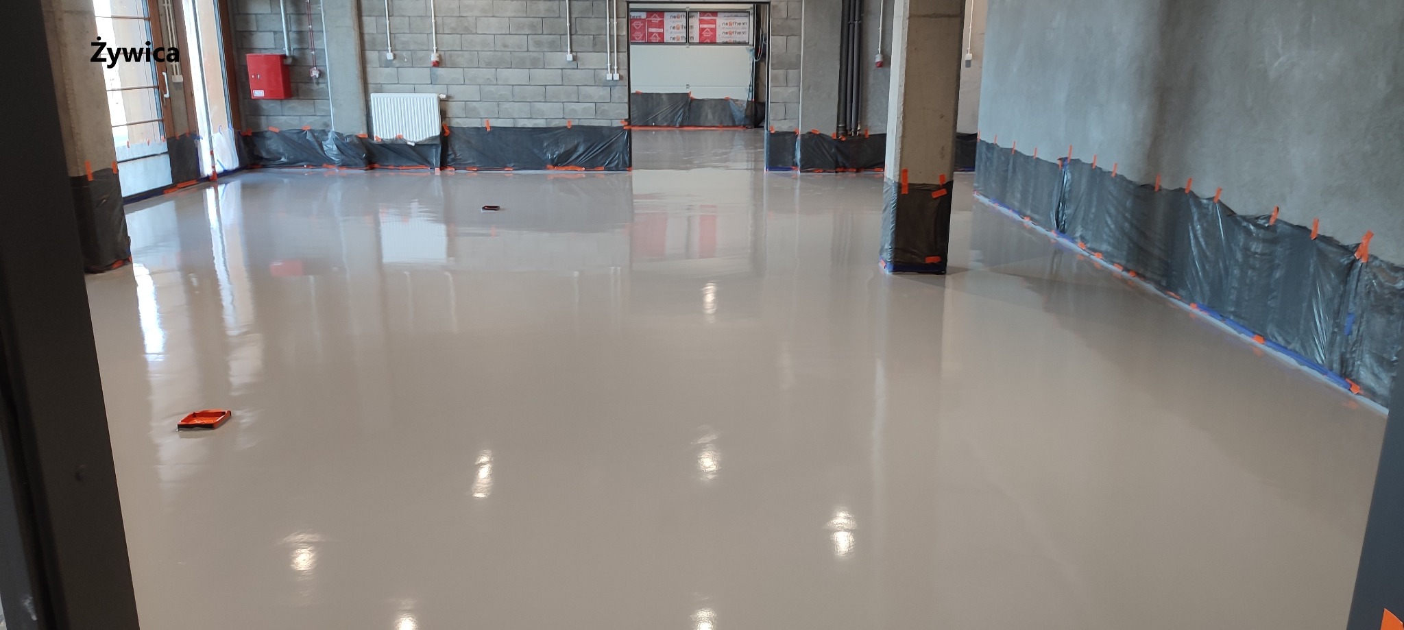 Świeżo wylana, beżowa posadzka żywiczna w hali przemysłowej, zabezpieczone ściany folią, widoczny wałek malarski na podłodze.