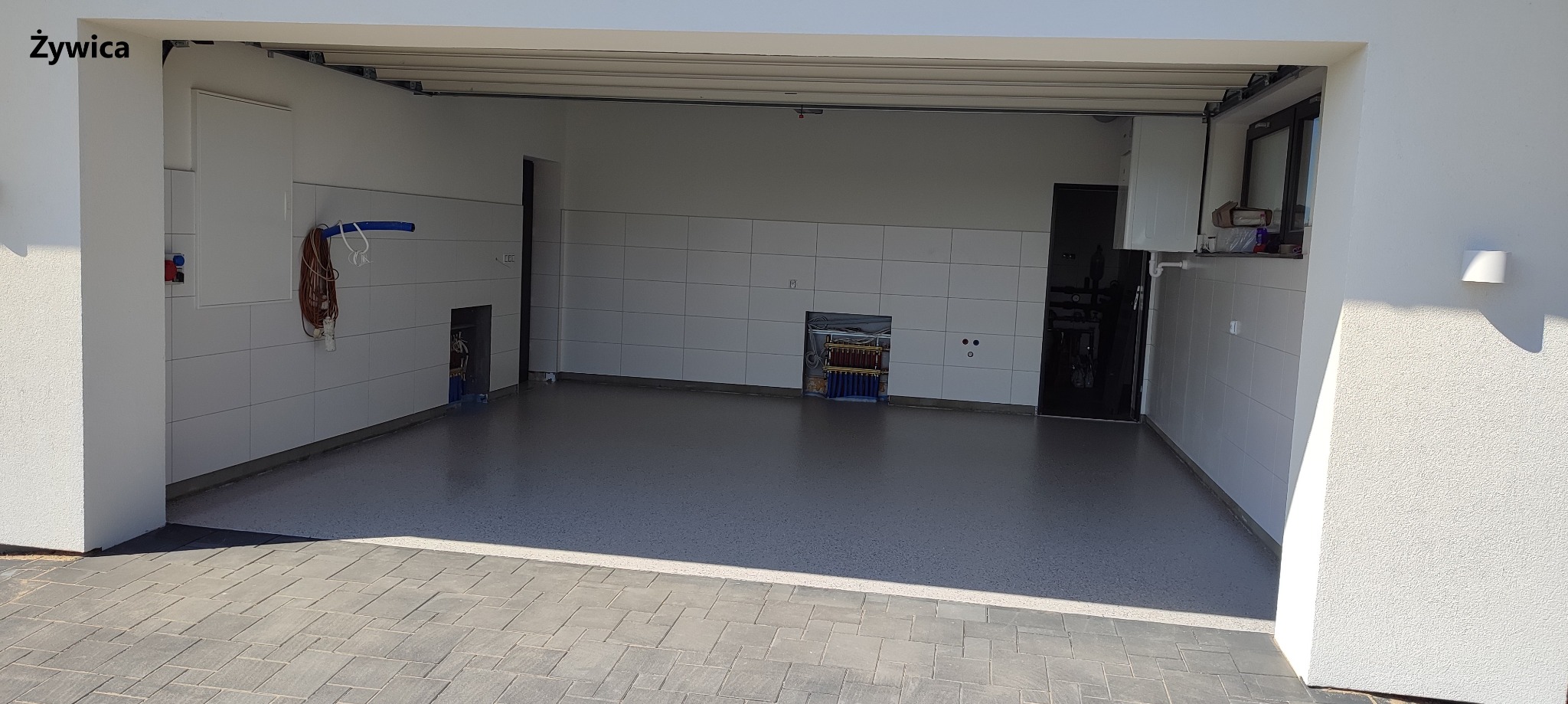 Wykończony garaż z szarą posadzką z delikatnym wzorem, białe płytki na ścianach, widoczne instalacje i osprzęt elektryczny, jasne wnętrze.