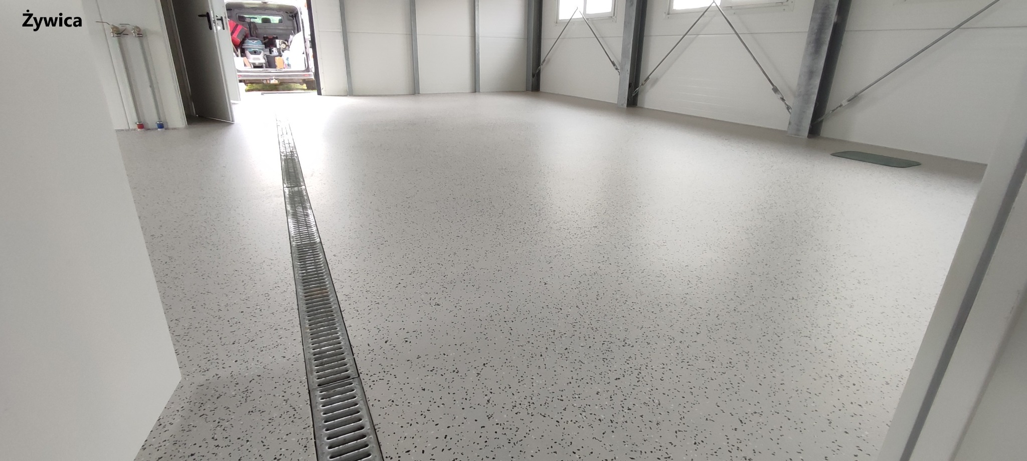 Jasnoszara, bezspoinowa posadzka z drobnymi, ciemnymi plamkami w hali magazynowej lub produkcyjnej. Widoczny liniowy odpływ podłogowy ze stali nierdzewnej oraz częściowo otwarty samochód dostawczy...