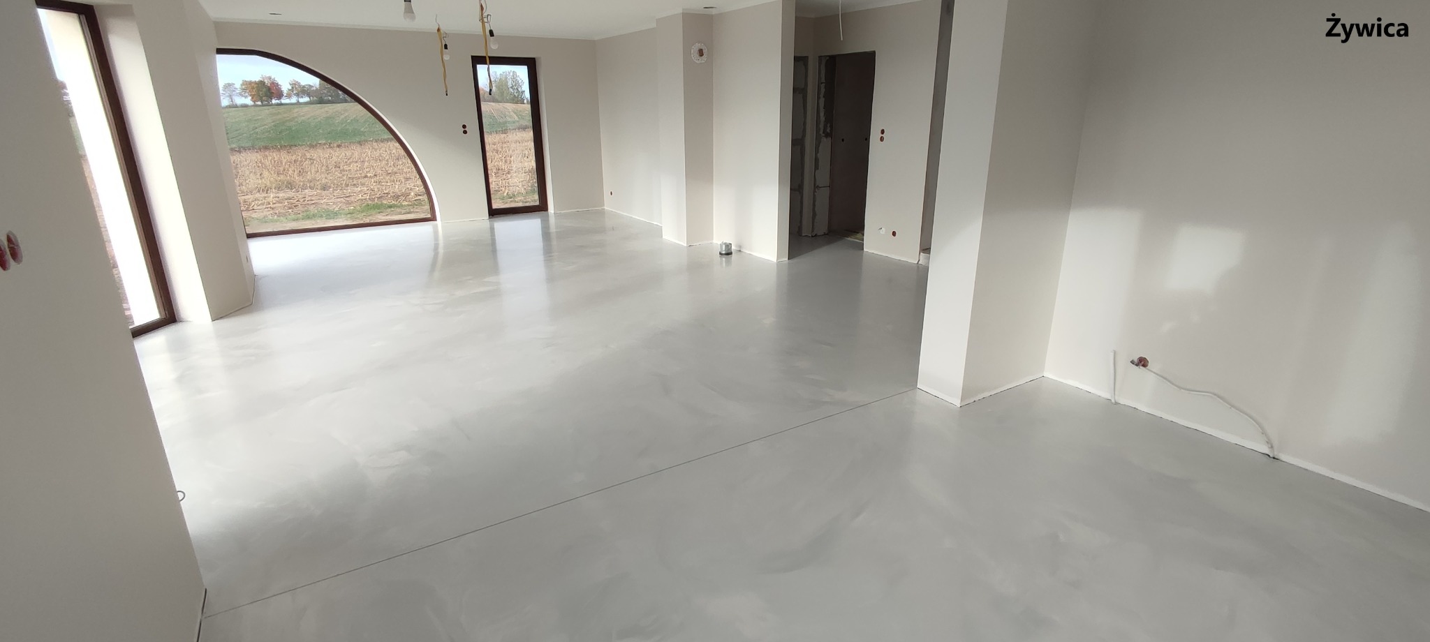 Jasnoszara, błyszcząca posadzka żywiczna w przestronnym wnętrzu z dużymi oknami, widok na jesienny krajobraz za oknem.