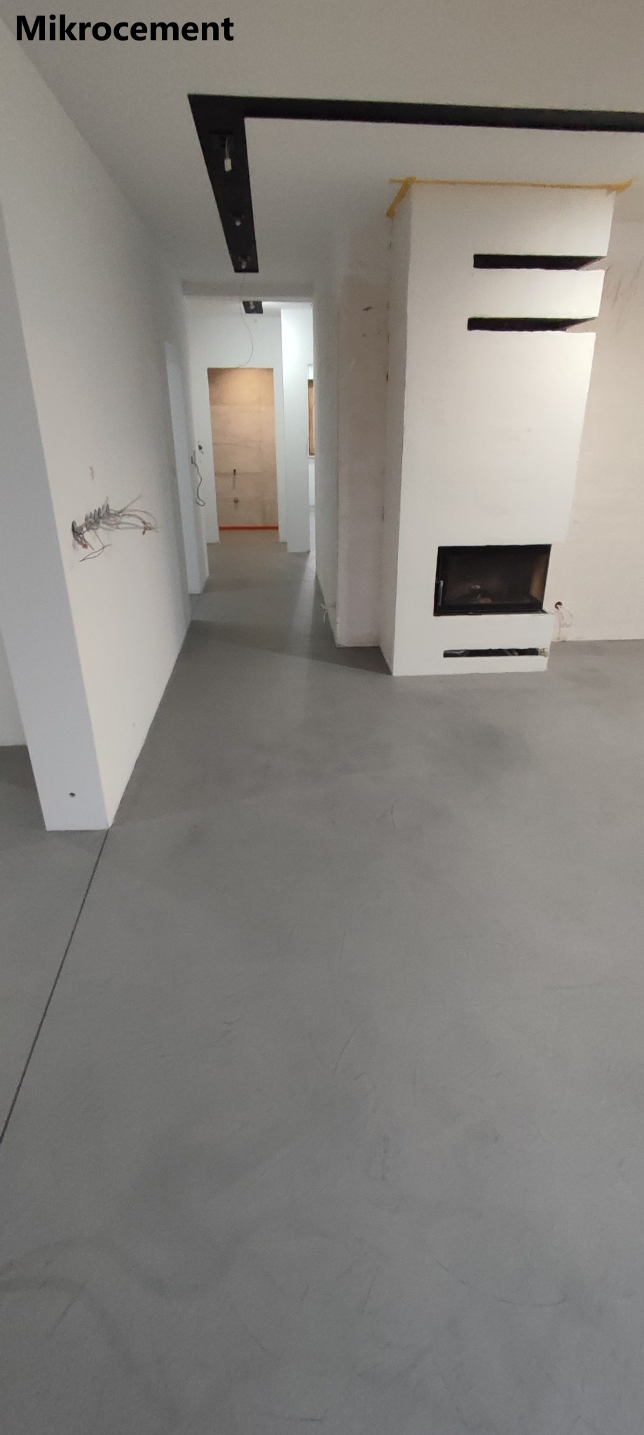 Szara, gładka posadzka w nowym wnętrzu z kominkiem; widoczne niedokończone ściany i wystające przewody elektryczne.