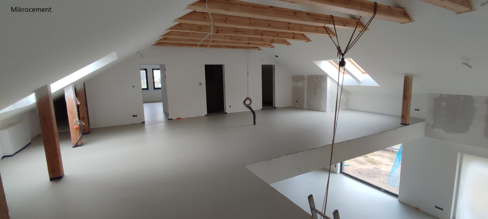 Jasnoszara, gładka podłoga w przestronnym, surowym wnętrzu poddasza z drewnianymi belkami stropowymi i pionowymi słupami, widoczne okno dachowe i otwory drzwiowe.