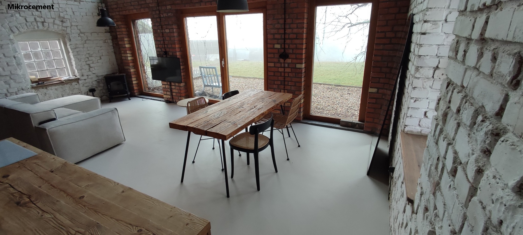 Jasnoszara podłoga w przestronnym wnętrzu z ceglanymi ścianami, drewnianym stołem i widokiem na ogród przez duże okna.