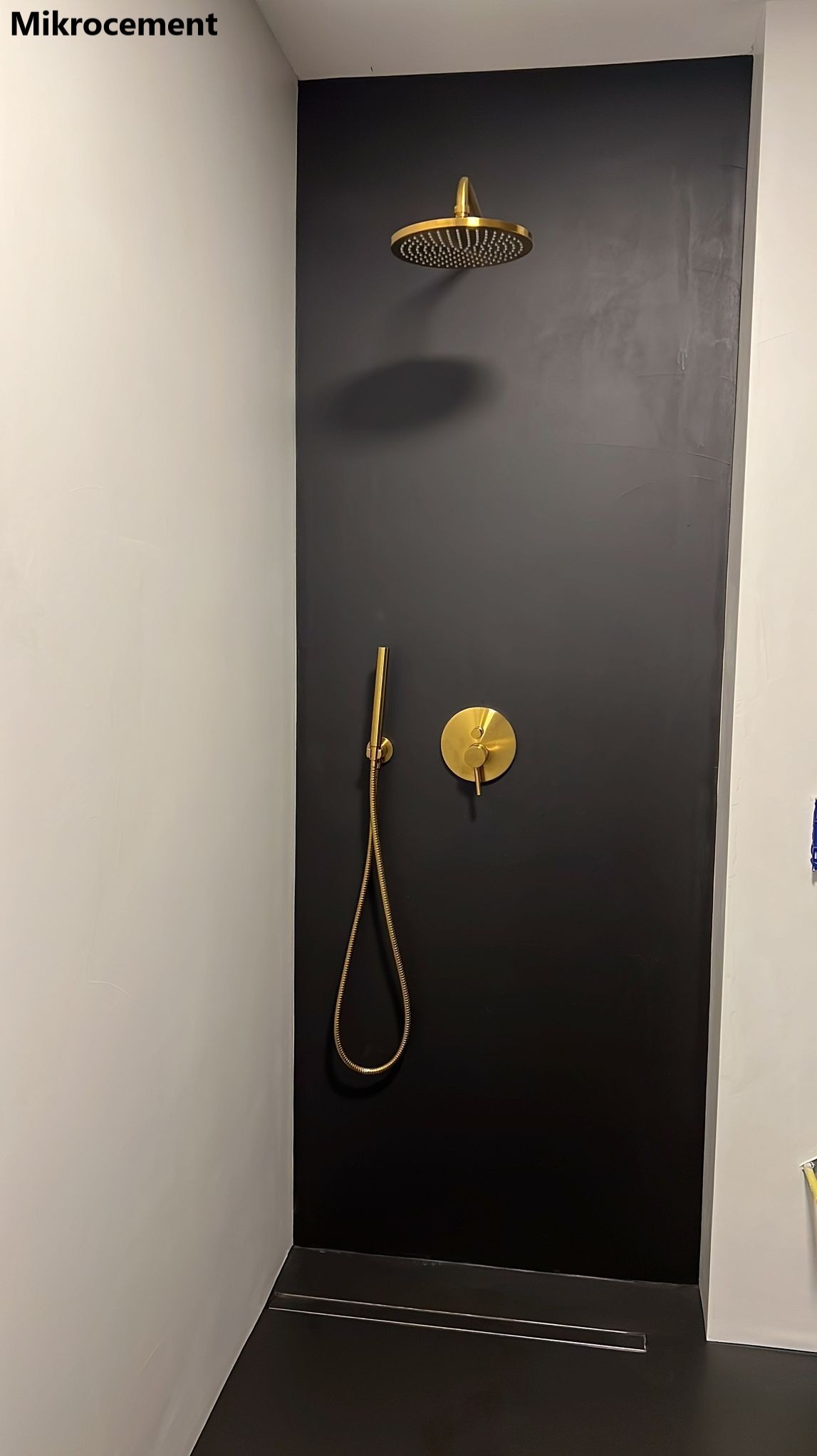 Czarna kabina prysznicowa z wykończeniem ścian i podłogi w ciemnym odcieniu. Złota deszczownica i bateria podtynkowa. Minimalistyczny odpływ liniowy.