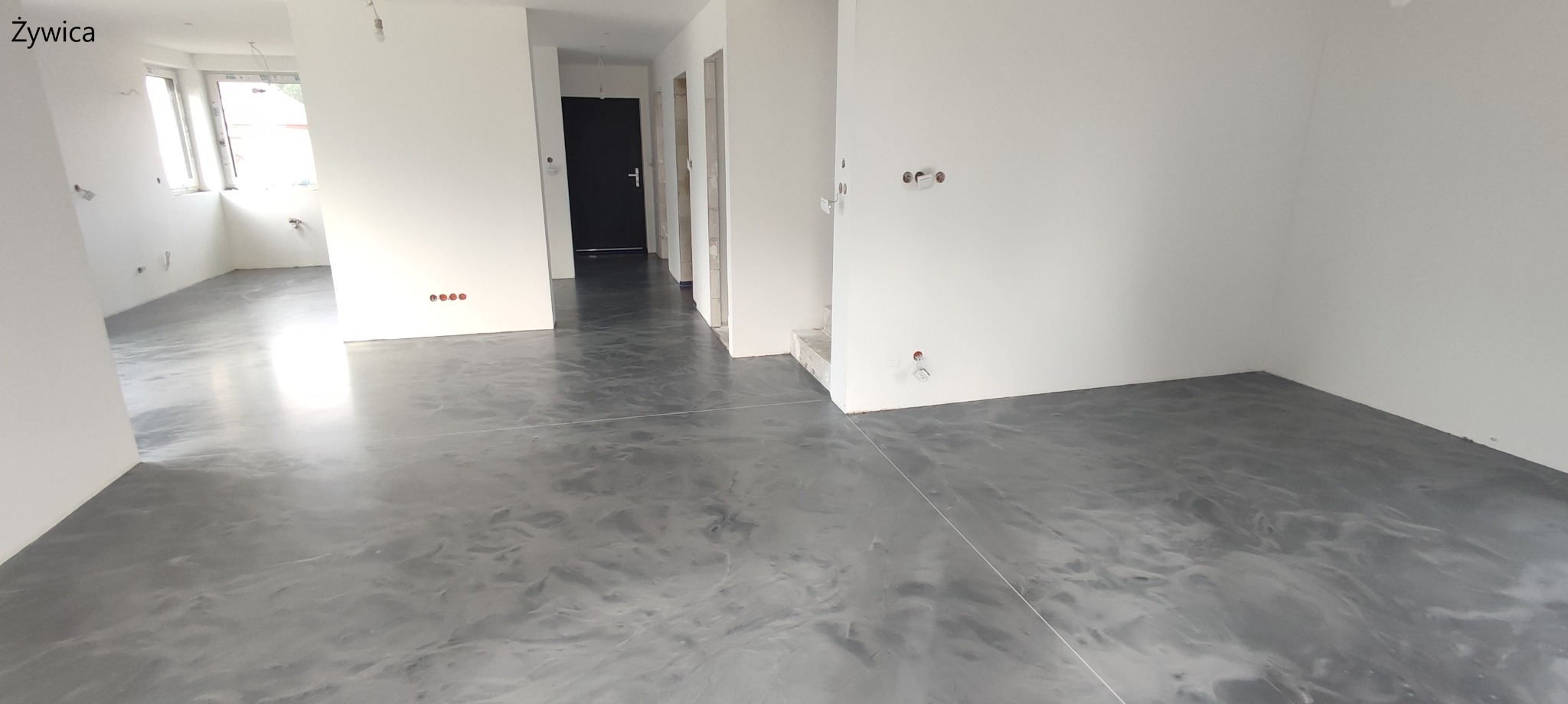 Szara, błyszcząca podłoga w surowym wnętrzu z widocznymi instalacjami elektrycznymi i otworami drzwiowymi.
