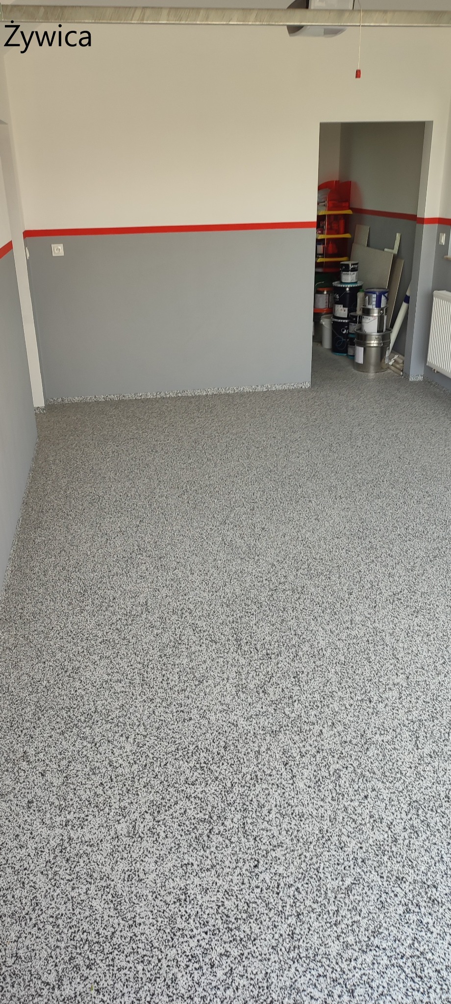 Szara posadzka żywiczna z drobnymi, czarnymi i białymi elementami w garażu lub pomieszczeniu gospodarczym, z widocznymi puszkami farb w tle.