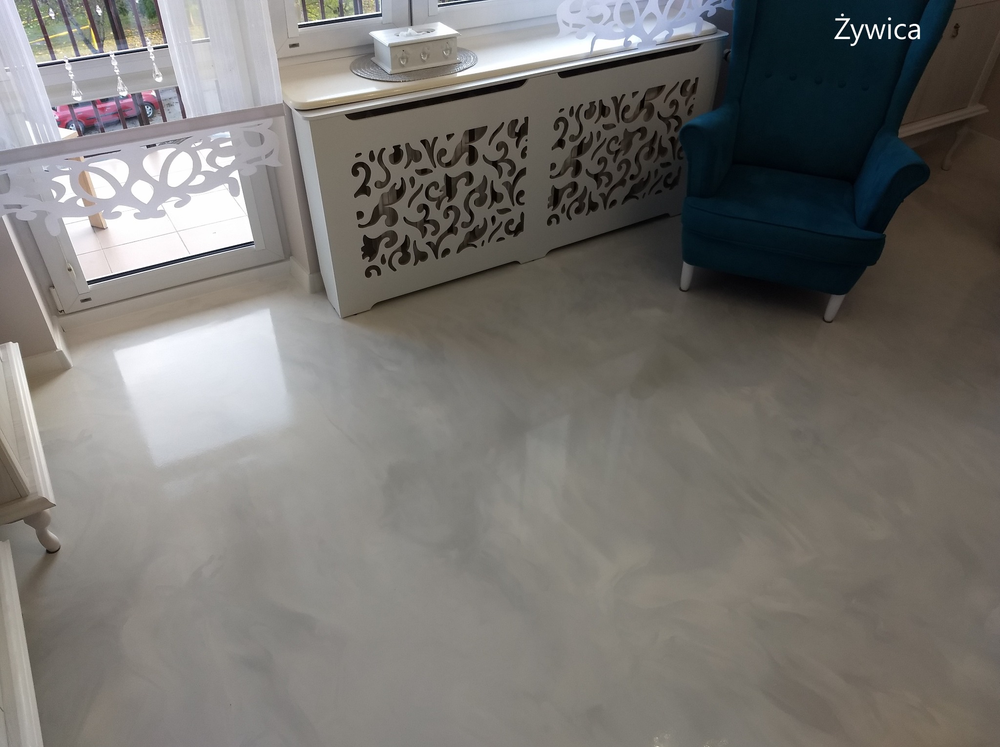 Jasnoszara, błyszcząca posadzka w pokoju z turkusowym fotelem i białym grzejnikiem z dekoracyjnym ażurem.