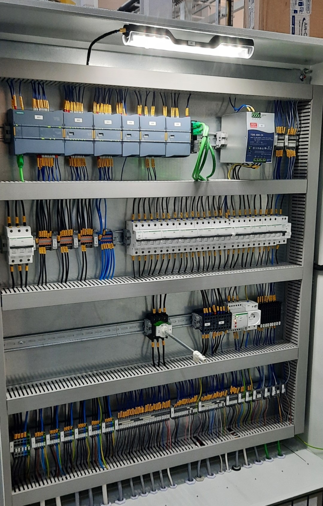 Rozdzielnica elektryczna z modułami sterującymi Siemens, zasilaczem Mean Well i licznymi przewodami podłączonymi do listew zaciskowych, oświetlona lampą LED wewnątrz szafy.
