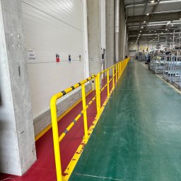 Stalweld - Żółta poręcz ochronna z czarnymi pasami, oddzielająca strefę czerwoną od zielonej podłogi w hali produkcyjnej, widok wzdłuż długiej, jasnej przestrzeni.