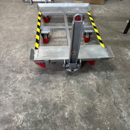 Stalweld - Wózek transportowy ze stali, z żółto-czarnymi pasami ostrzegawczymi, na czerwonych kółkach, stojący na betonowej posadzce w warsztacie.