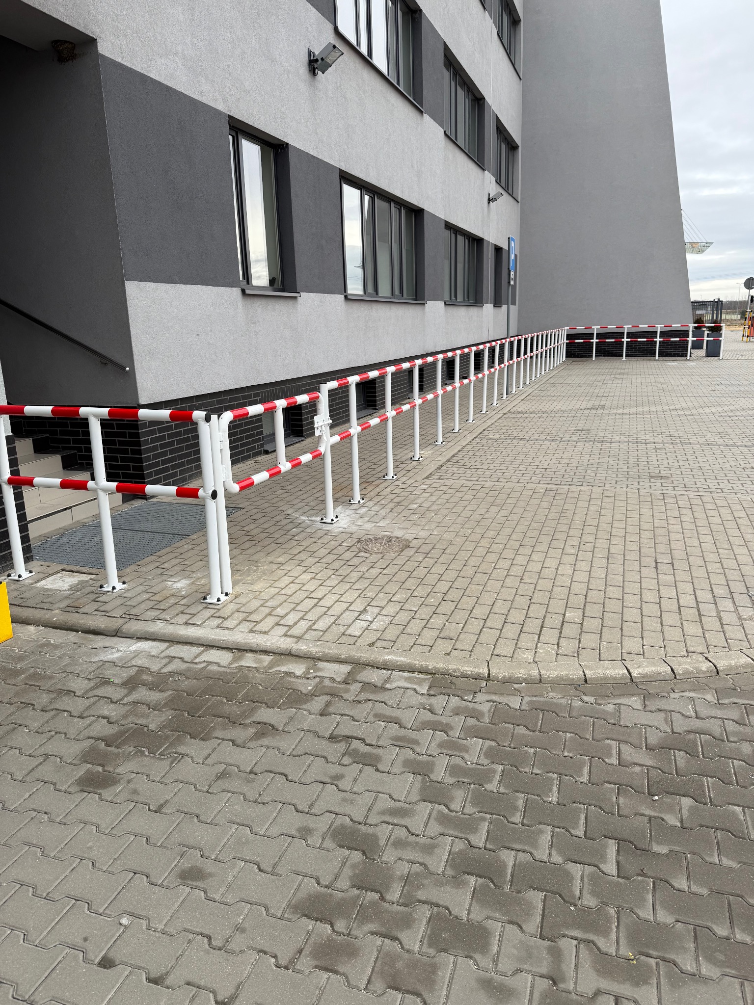 Białe poręcze z czerwonymi pasami zabezpieczające podjazd dla niepełnosprawnych przy nowoczesnym budynku biurowym z szarą elewacją.