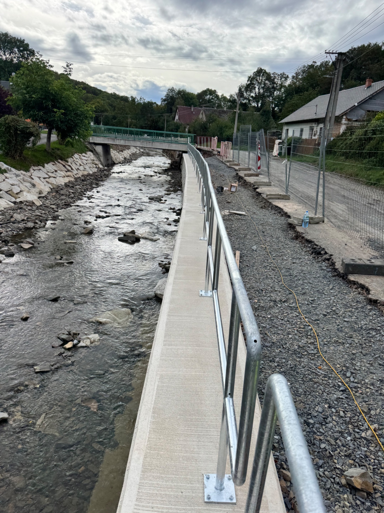 Metalowa poręcz zabezpieczająca brzeg betonowego koryta rzeki w krajobrazie wiejskim z widocznym mostem w tle.