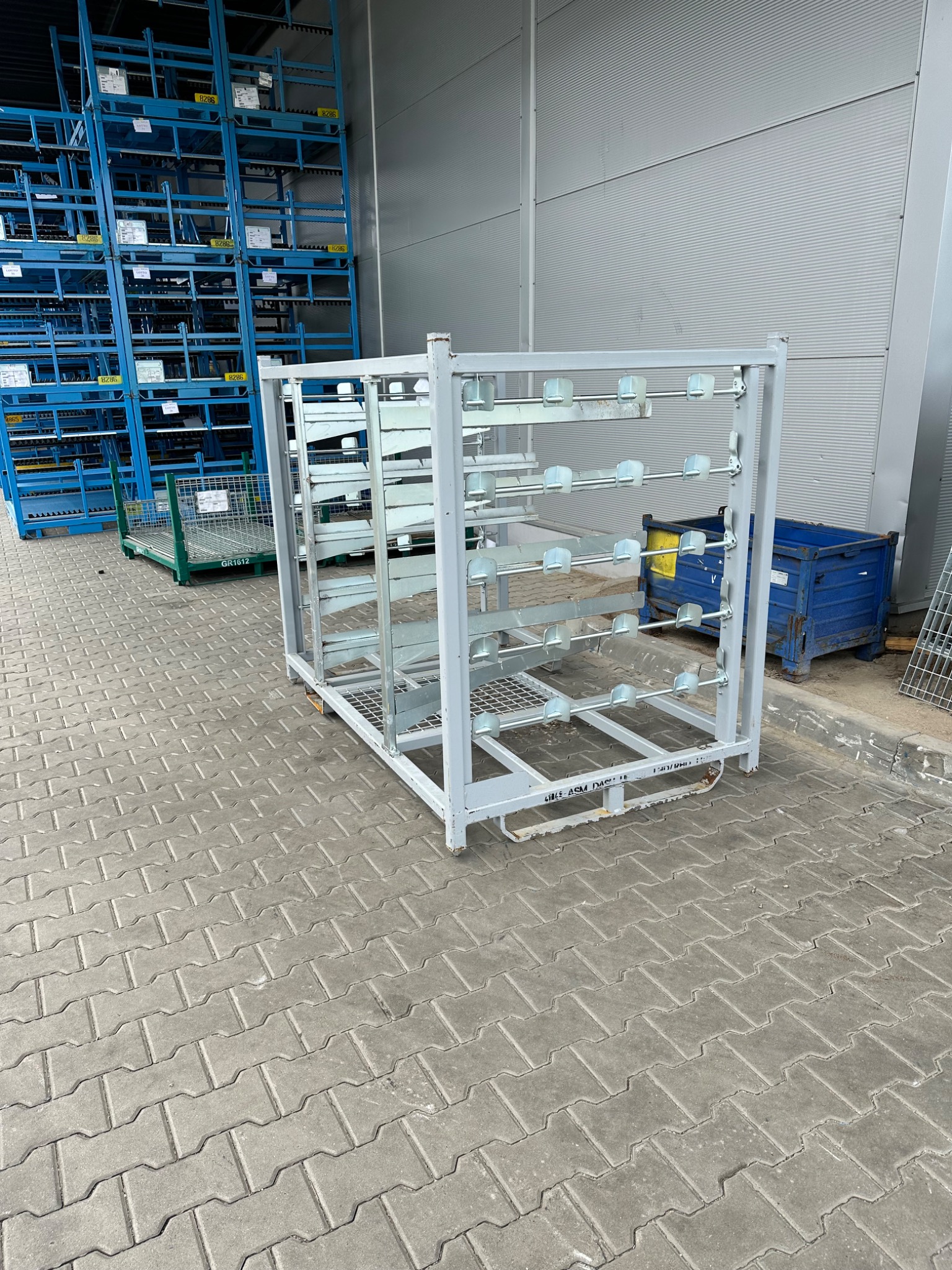 Metalowy, pomalowany na szaro, wózek transportowy z uchwytami, stojący na betonowej kostce brukowej, w tle magazyn z regałami.