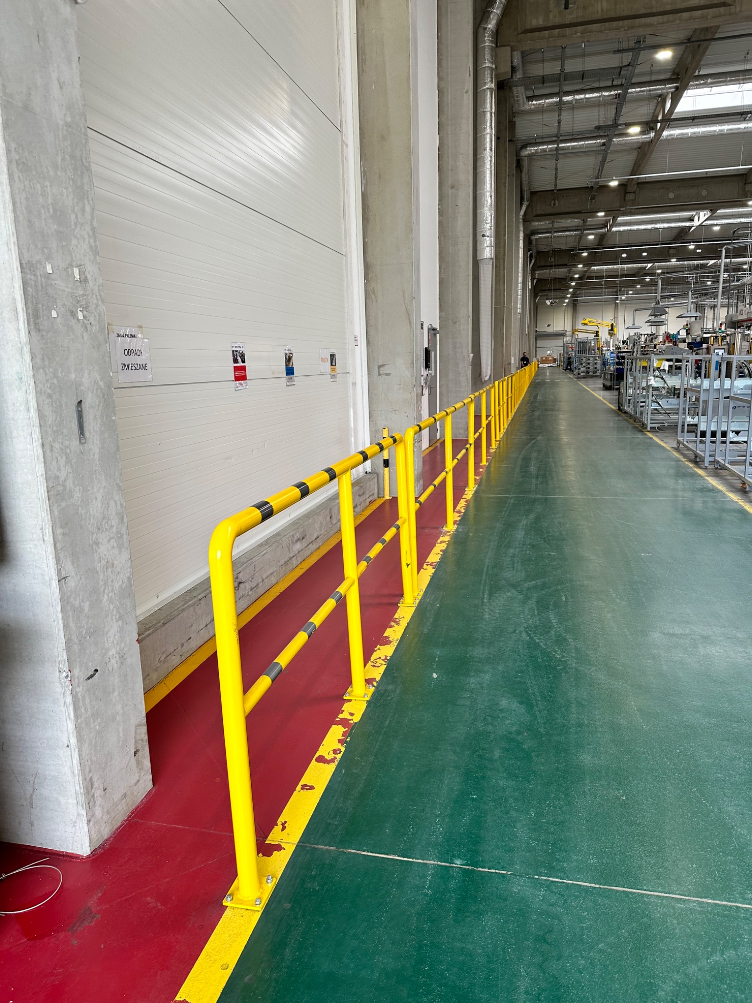 Żółta poręcz ochronna z czarnymi pasami, oddzielająca strefę czerwoną od zielonej podłogi w hali produkcyjnej, widok wzdłuż długiej, jasnej przestrzeni.