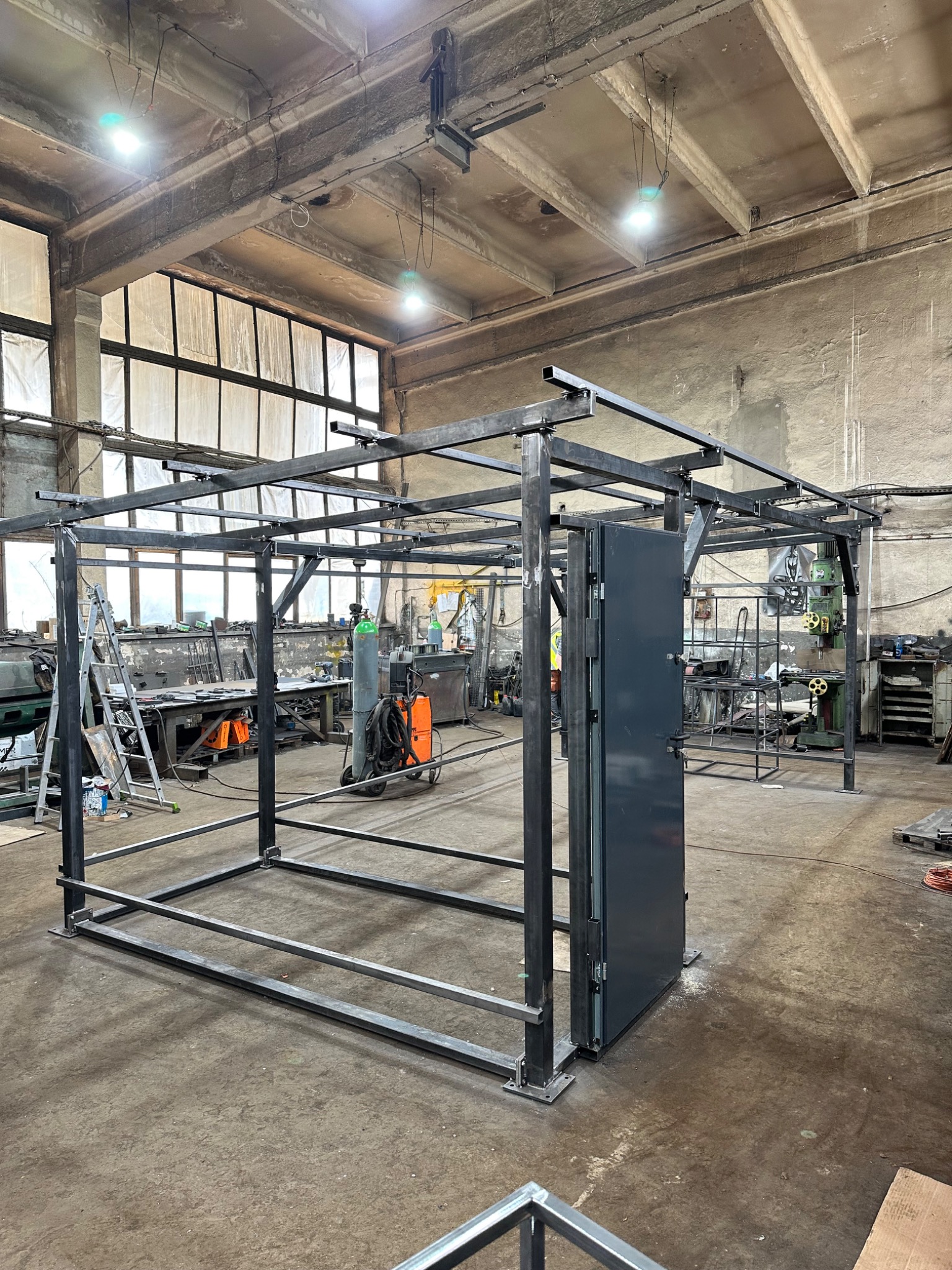 Konstrukcja metalowa z drzwiami w hali produkcyjnej, widoczny spawarki, butla z gazem i drabina.