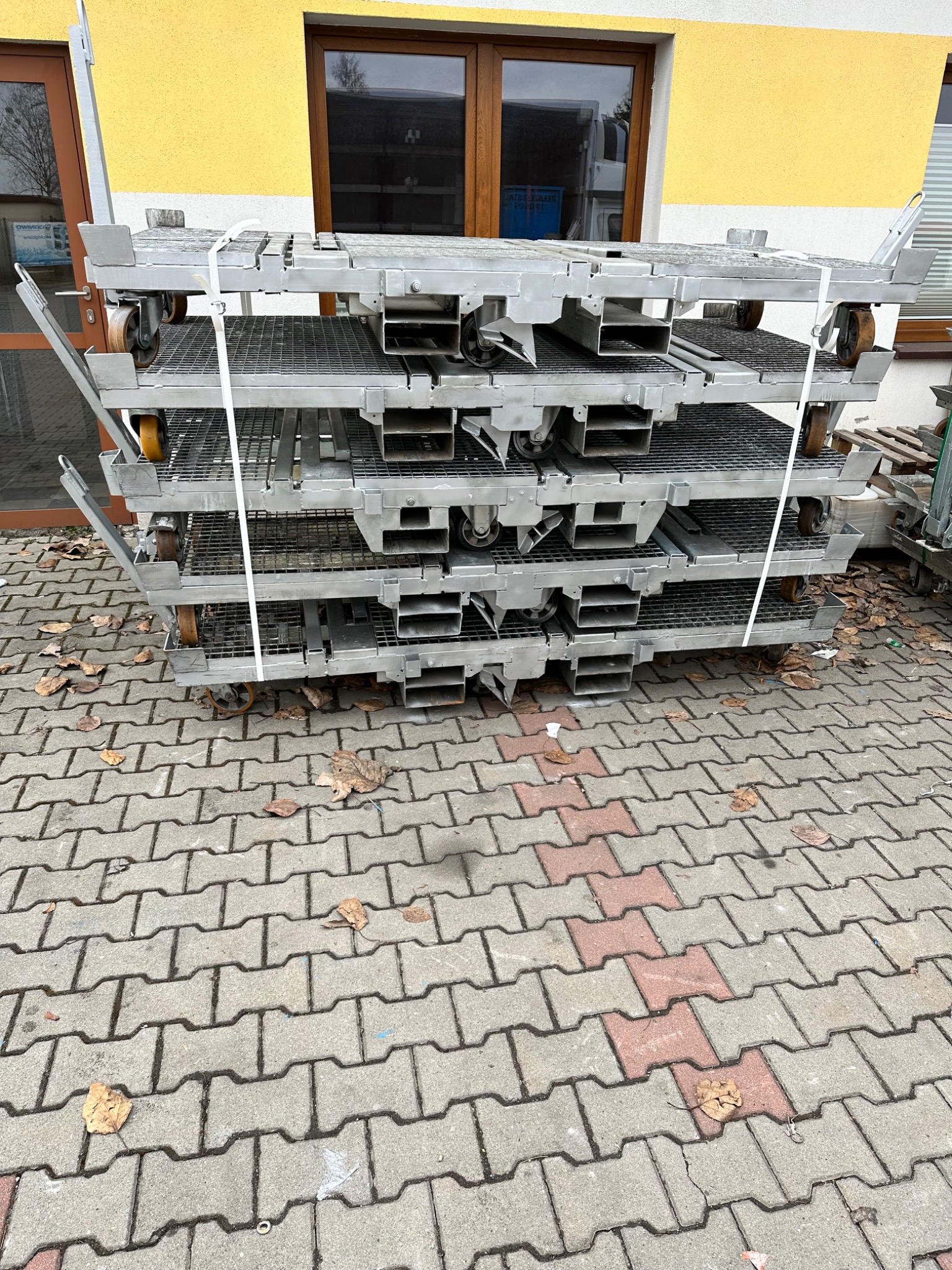 Stos metalowych platform transportowych na kółkach, zabezpieczonych białym sznurkiem, na tle kostki brukowej i ściany budynku z oknem.