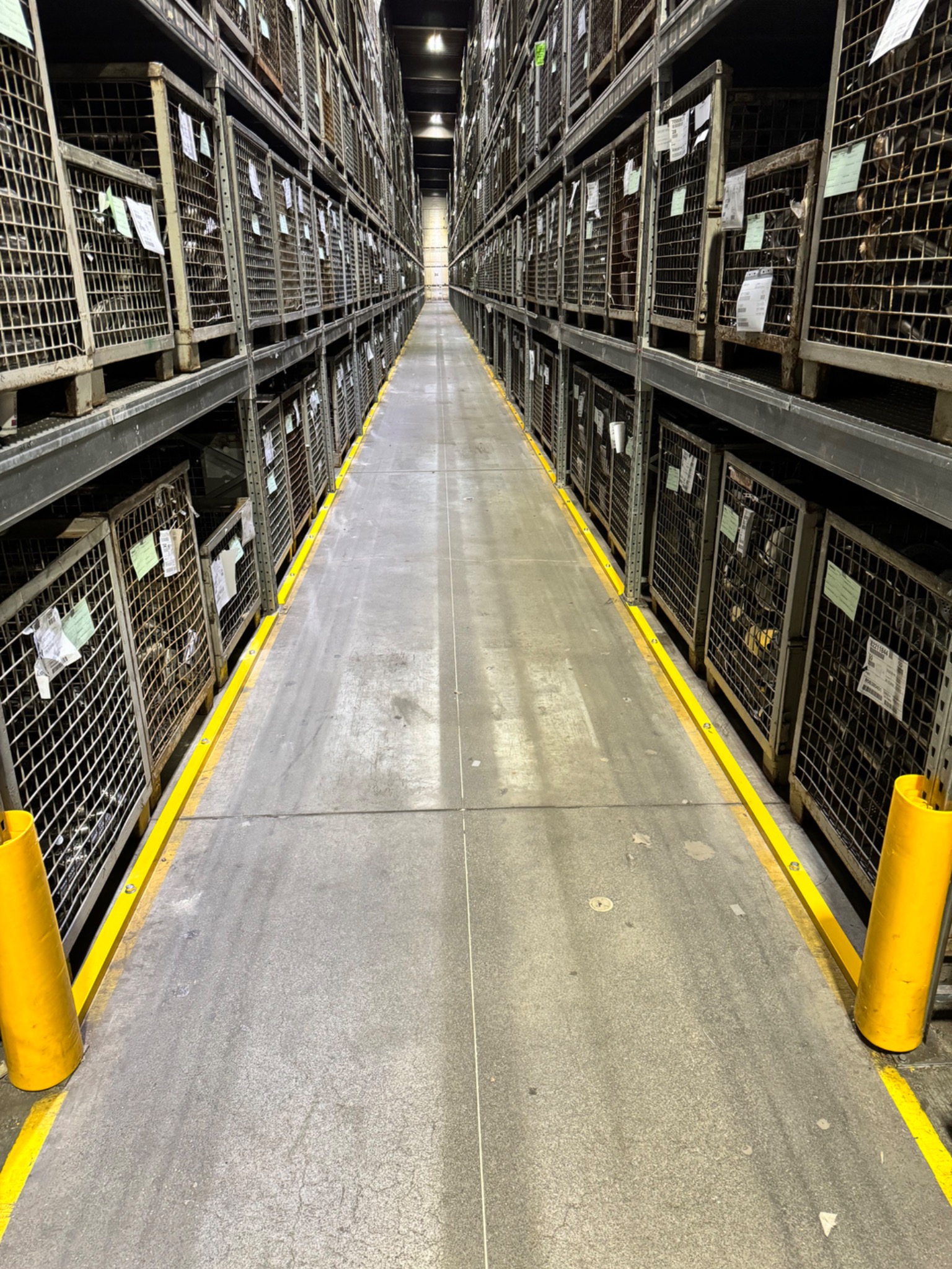 Długi korytarz magazynowy z metalowymi regałami wypełnionymi metalowymi koszami z siatki, zabezpieczony żółtymi barierami.